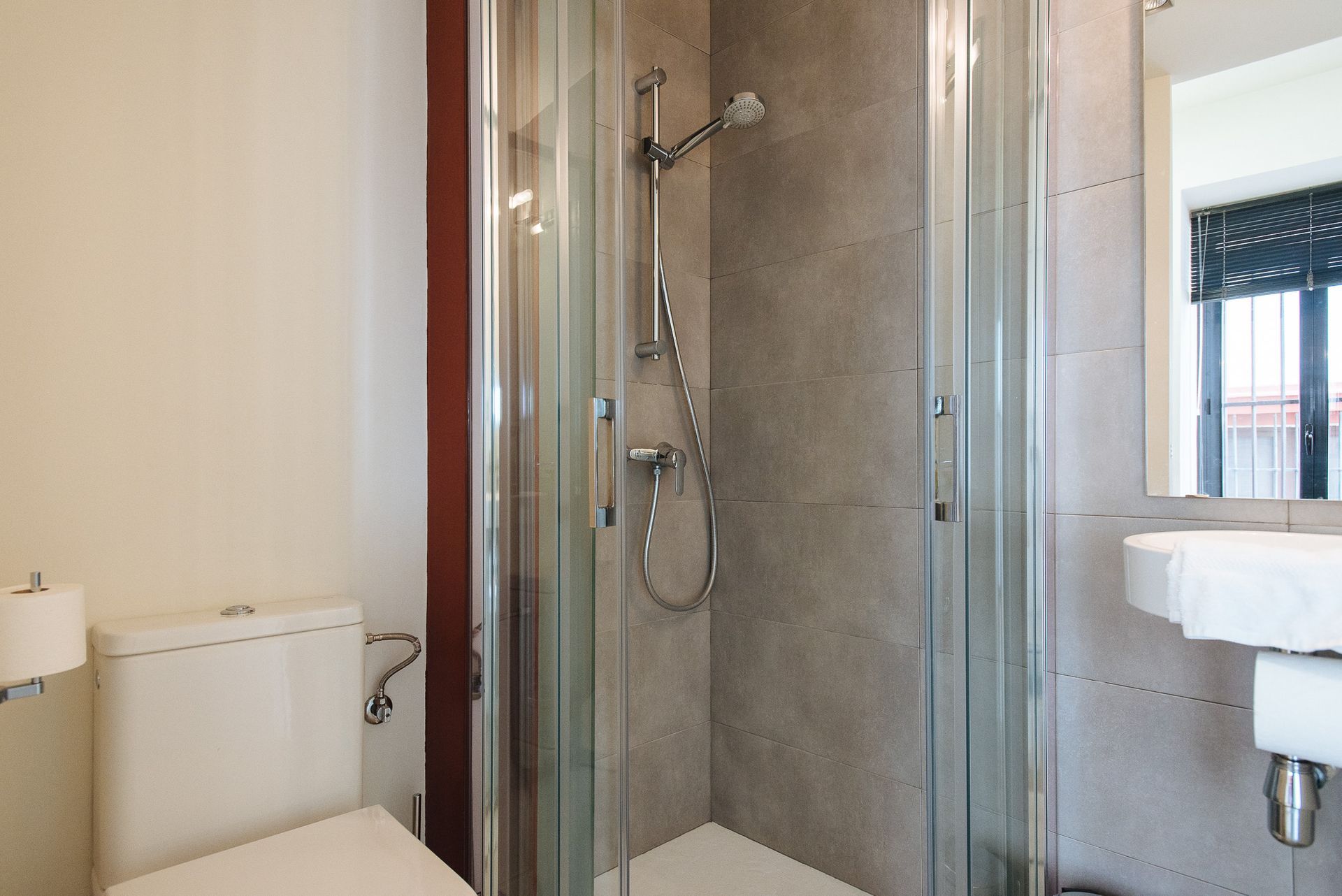 Bathroom with a glass shower, toilet, and a sink with a mirror above. Gray tile and beige walls.
