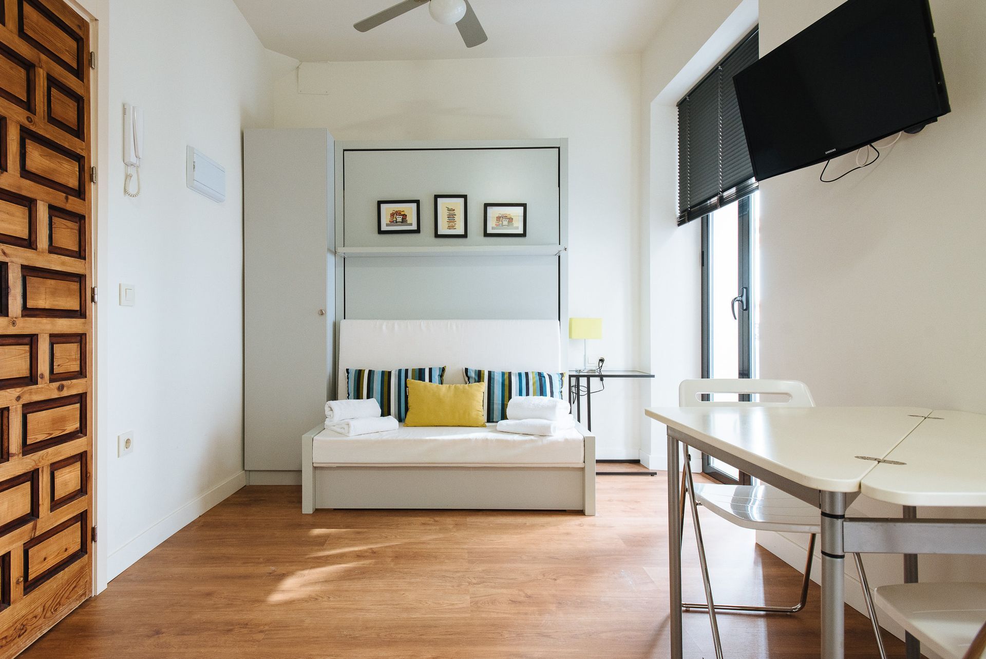 Small apartment interior: wooden floor, fold-down bed, small table, TV on wall, wooden door.