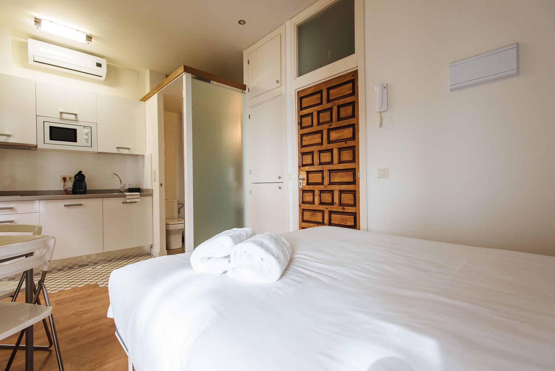 Small studio apartment with a bed, kitchenette, and wooden door. White towels on the bed.