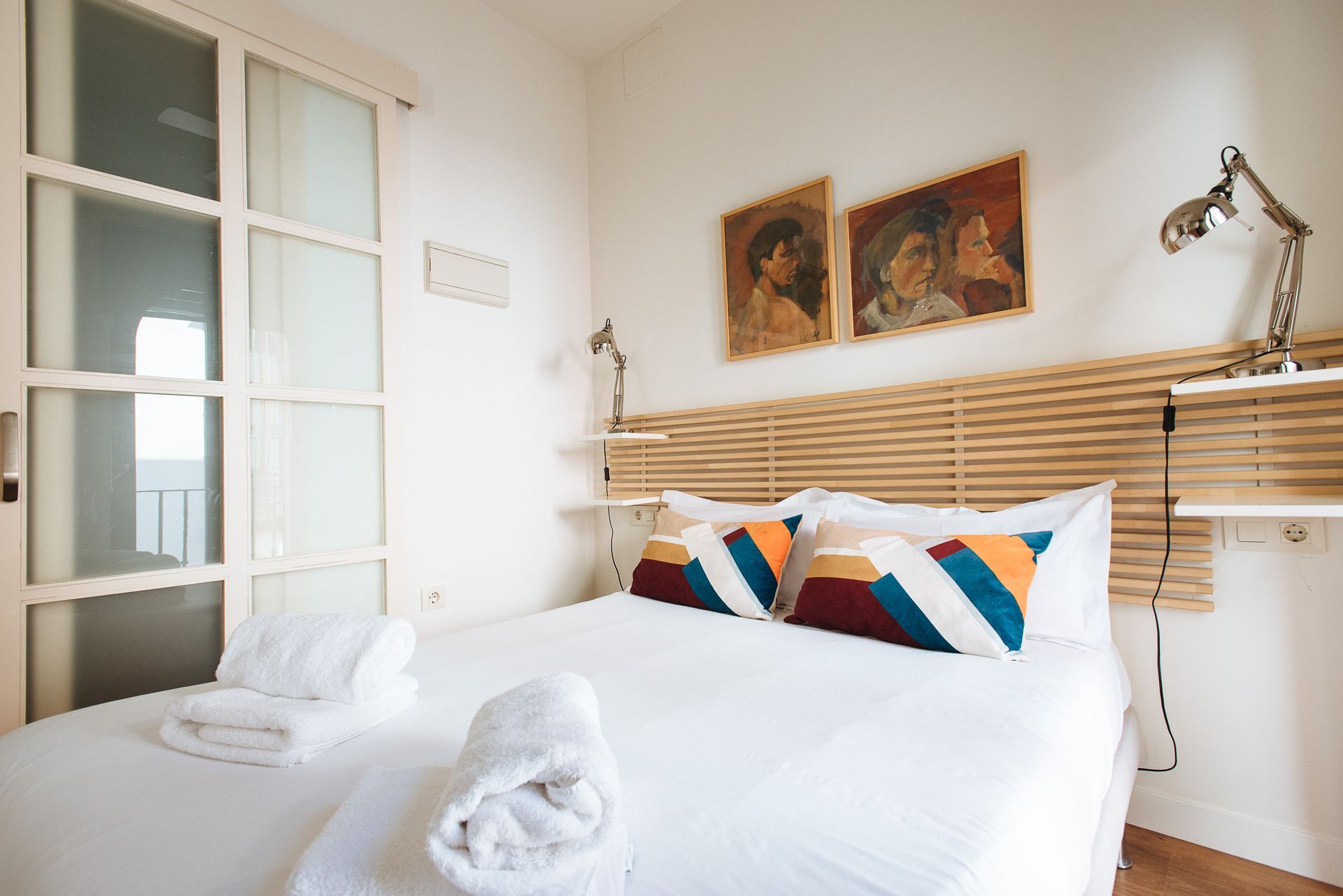 Bedroom with white walls, bed with colorful pillows, artwork above the headboard, and sliding glass door.