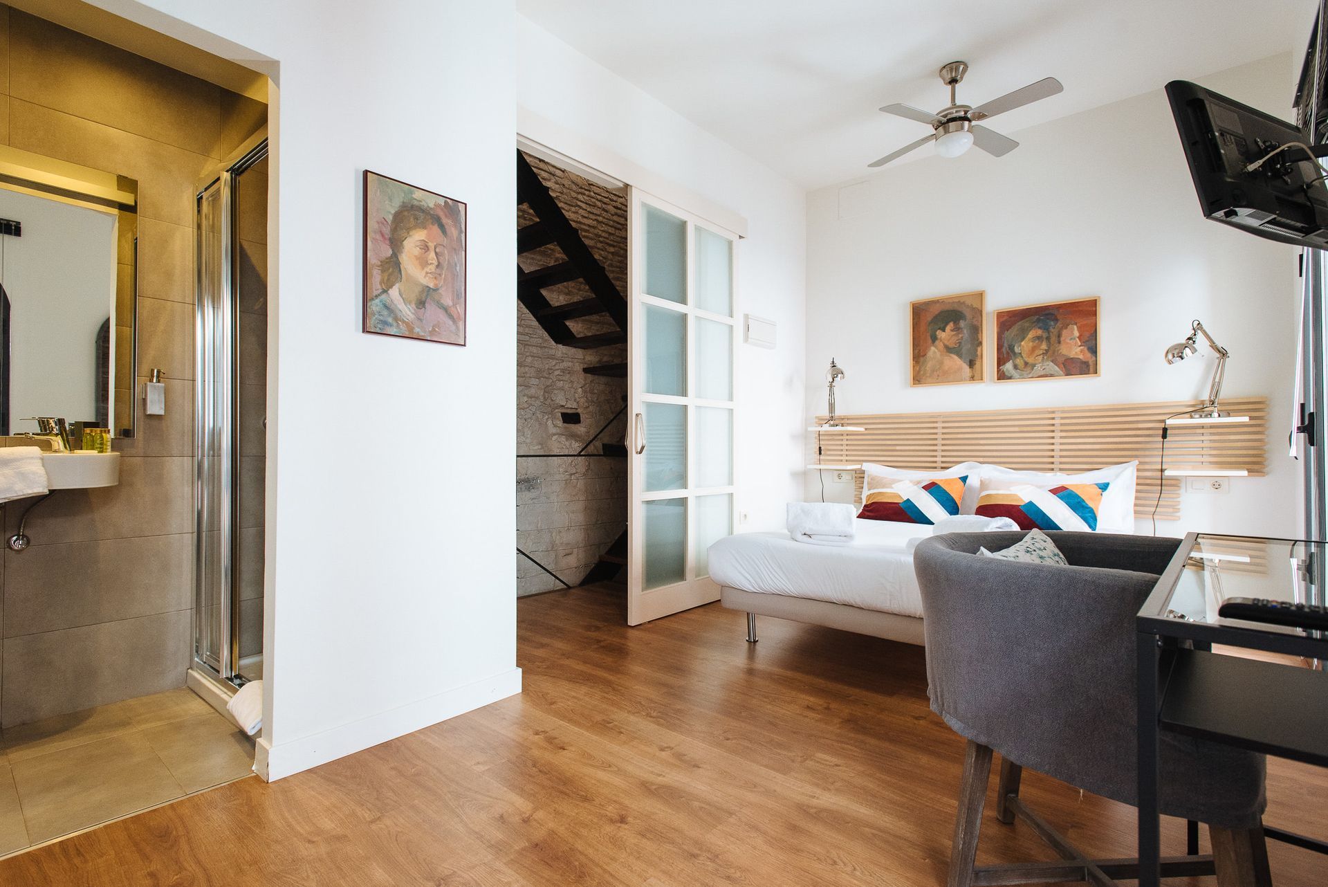 Bedroom with a bed, desk, bathroom, and artwork. Natural light and wooden floors.