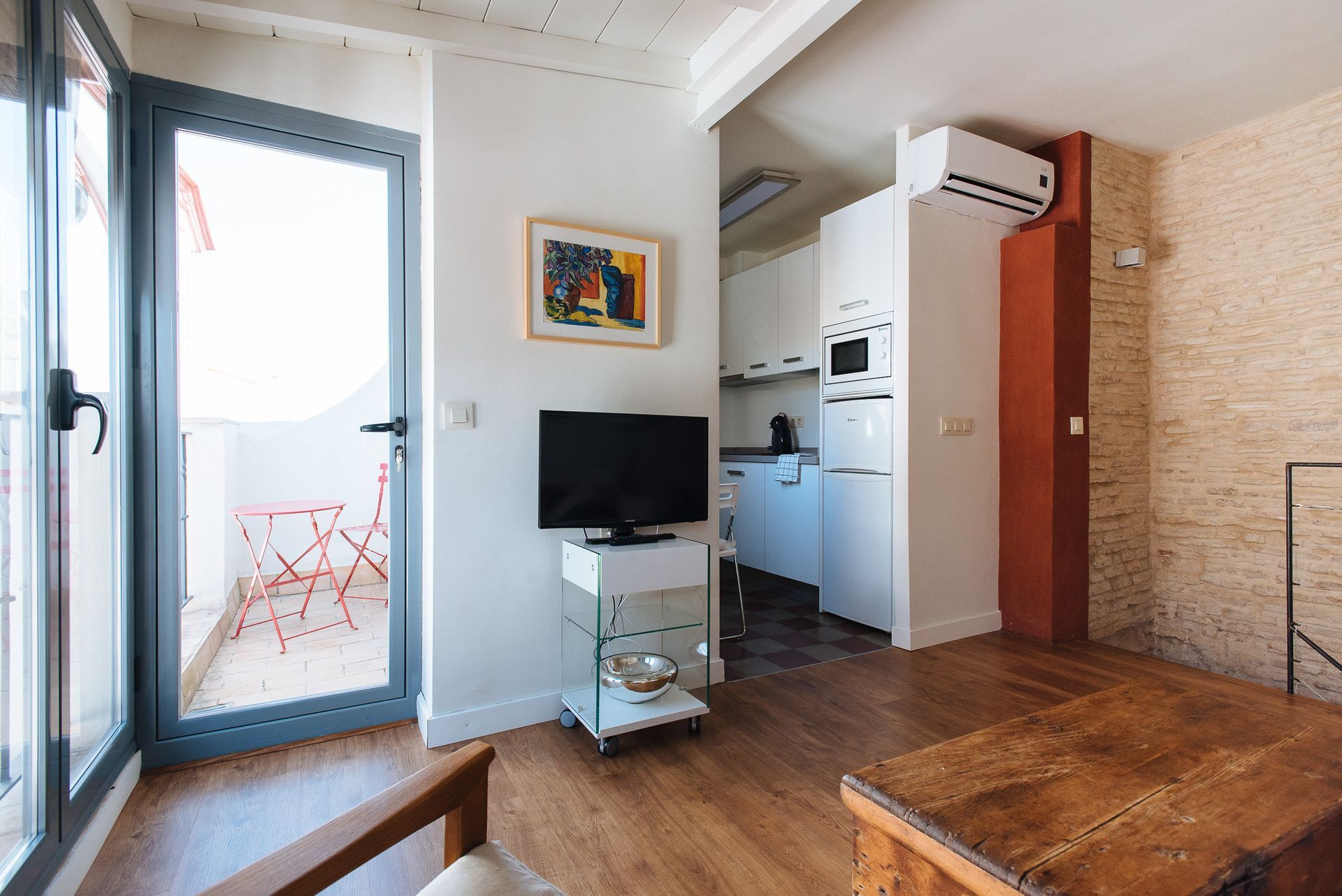 Small studio apartment with balcony, kitchen, and television. White walls, wooden floors, and red accents.