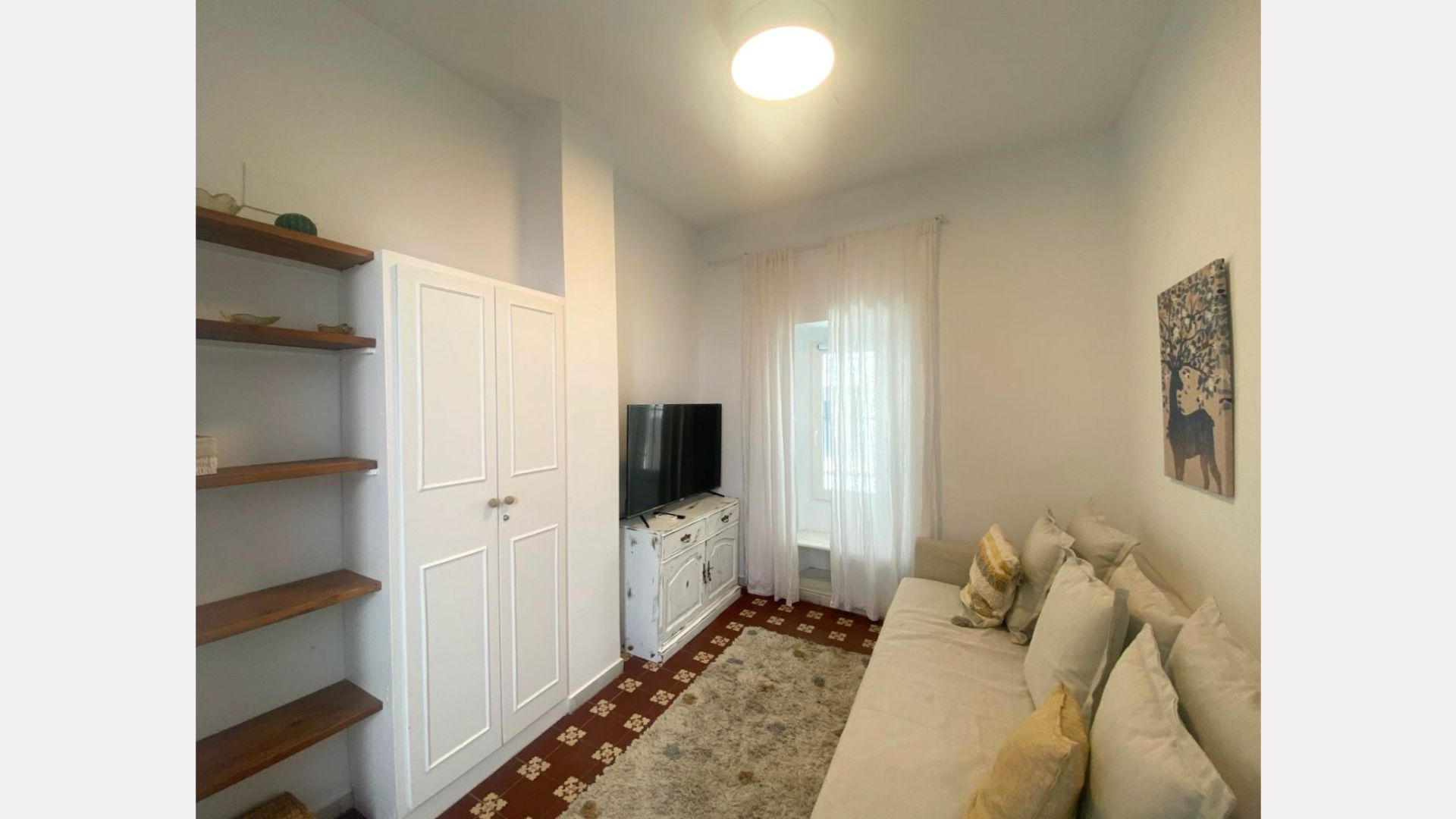 Small, bright room with white furniture, TV, and couch; brown shelves and patterned floor.