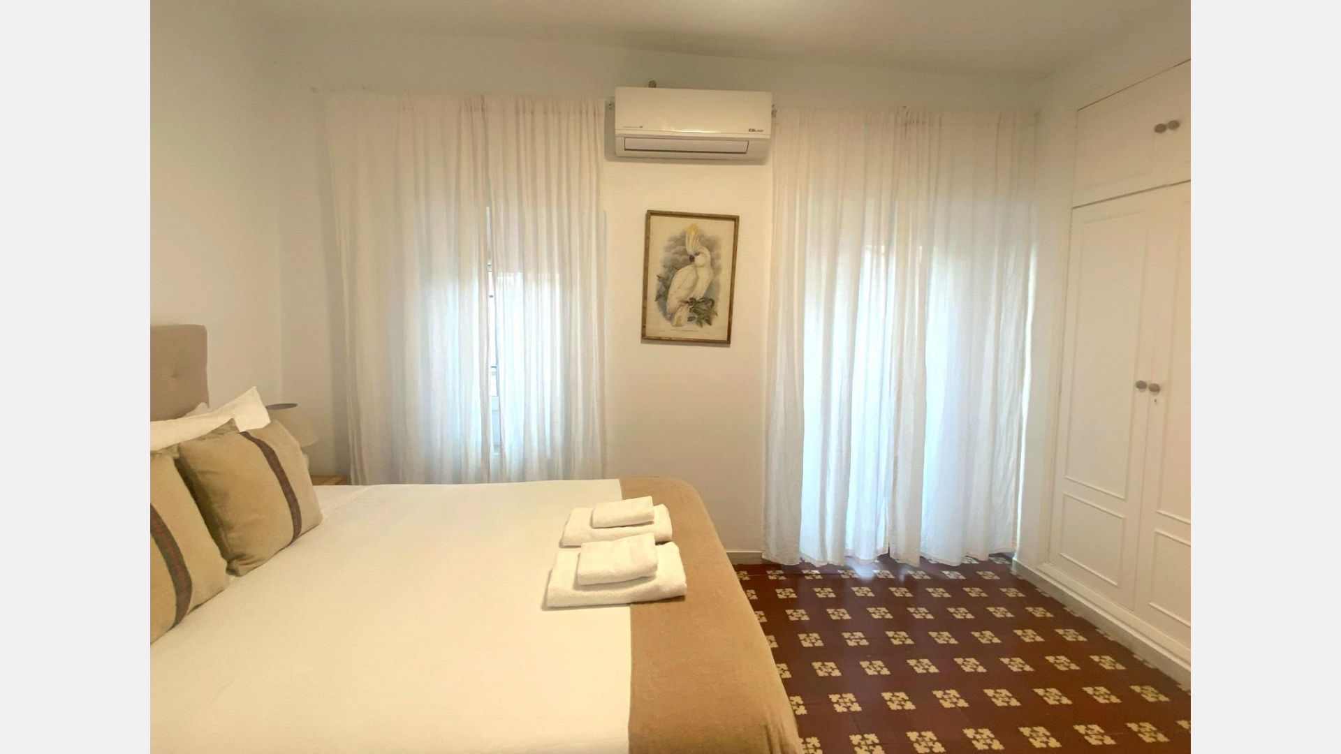 Bedroom with a large bed, white curtains, and patterned floor. Air conditioner above the bed.