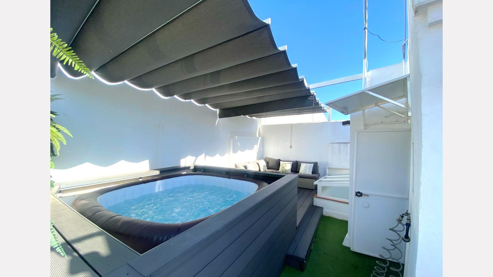 Outdoor hot tub with gray canopy, deck, white walls, and blue sky.