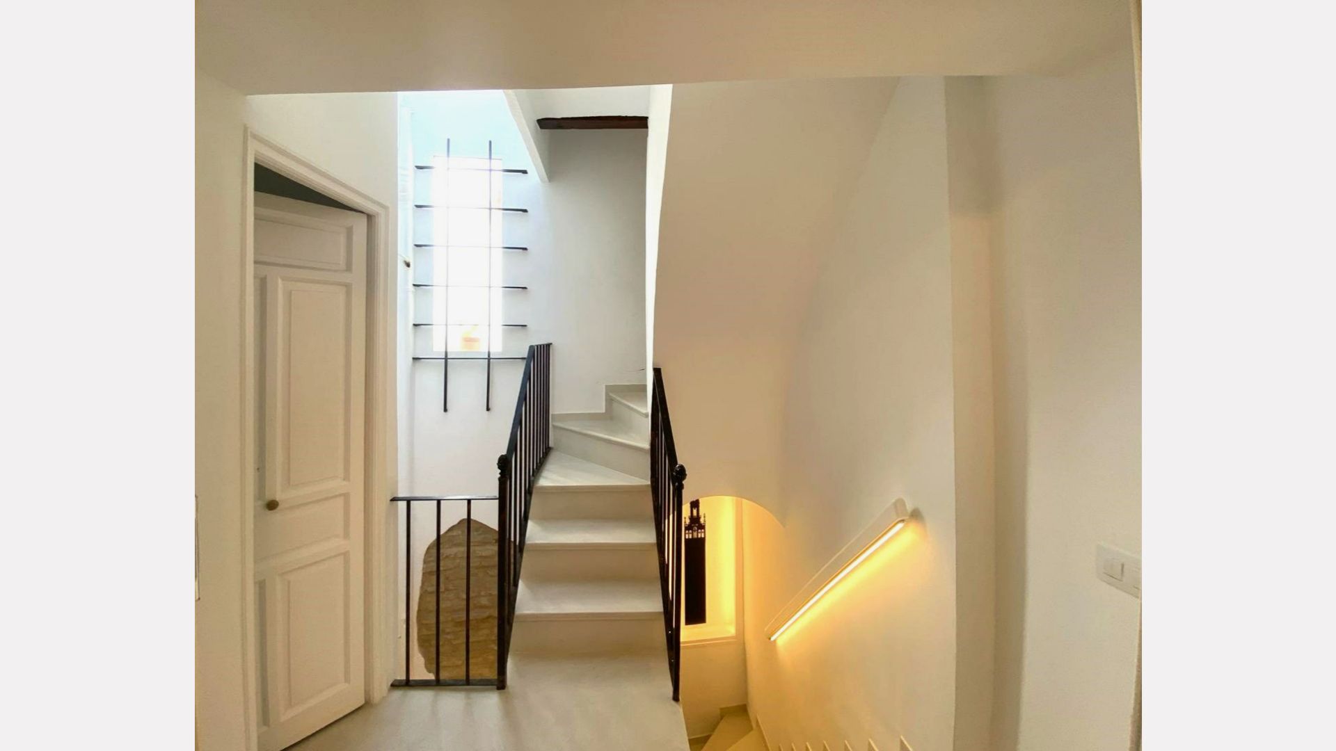 Interior hallway with staircase, white walls, and a light-up handrail. A door is on the left.