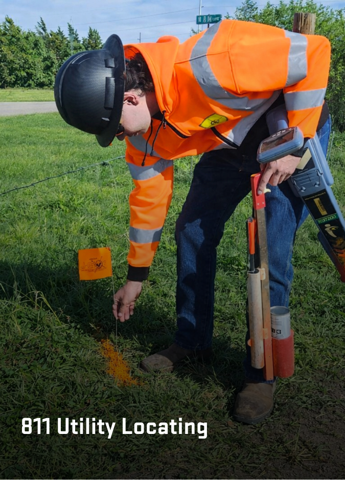 A person in a high-visibility orange jacket and black hard hat marks the ground with orange paint for utility locating.
