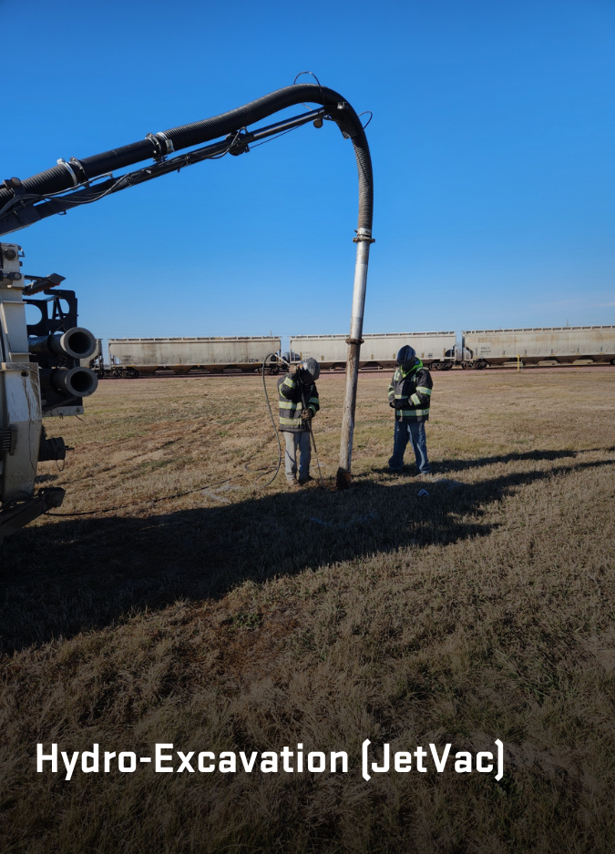 Two workers use a truck-mounted hydro-excavator to dig into a grassy field with train cars in the distance.