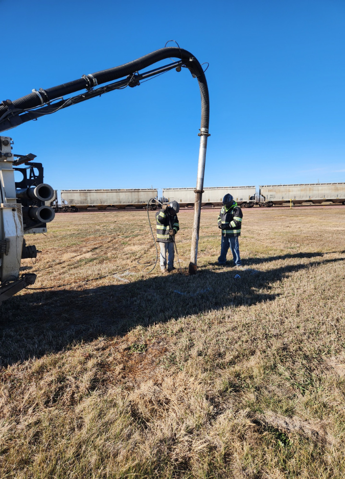 Two workers in high-visibility gear use a vacuum truck boom to excavate in a field near a train.