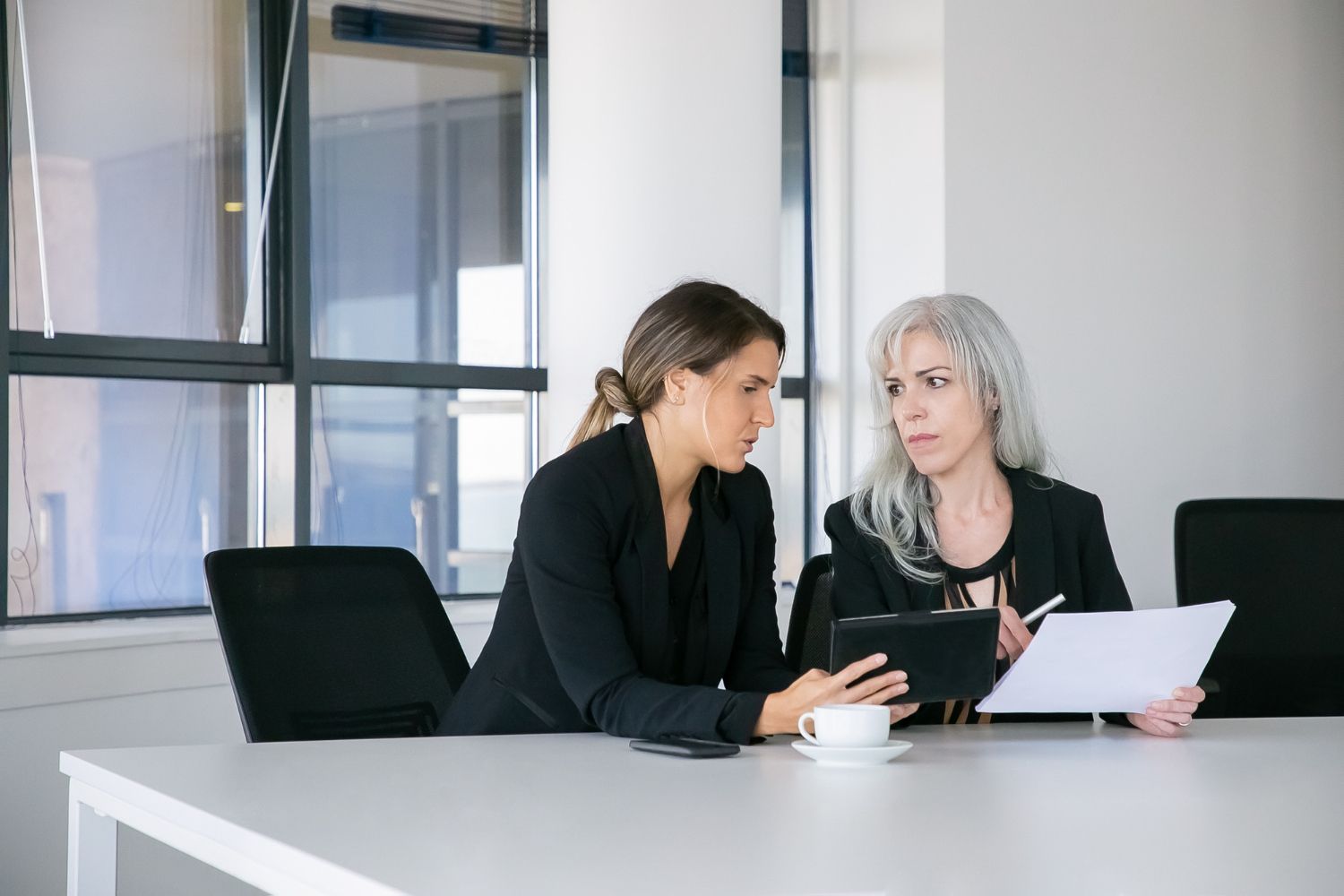 Two people in a meeting room review documents beside a tablet at a white table.