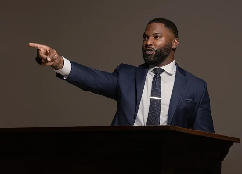 Man in navy suit pointing while speaking at a podium on a stage