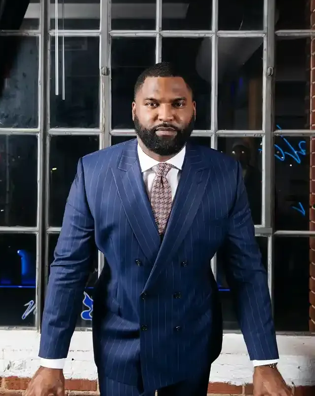 Man in a blue pinstripe suit stands indoors before a windowed wall.