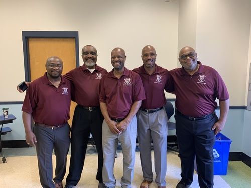 Five men in maroon shirts pose indoors, smiling, some with arms around each other.