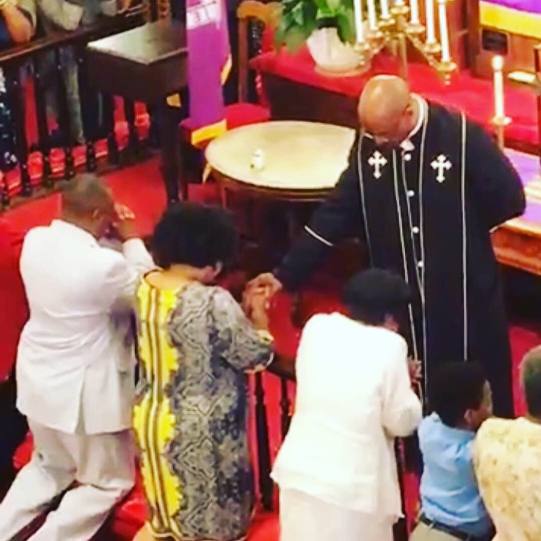 People kneeling in prayer as a robed figure touches their heads, inside a church.
