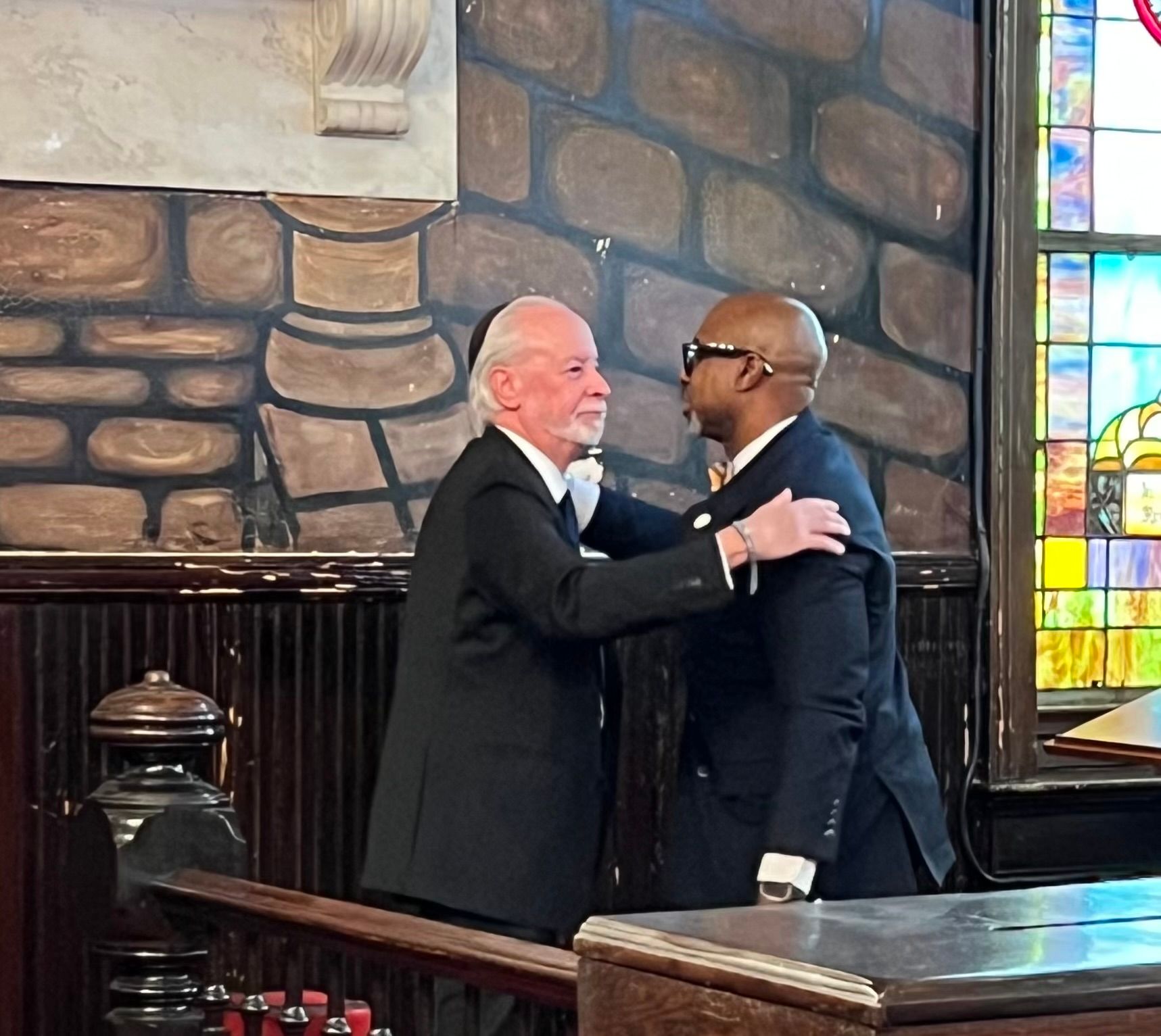Rabbi Myers and Rev. Manning embrace inside a church, stained glass window visible.