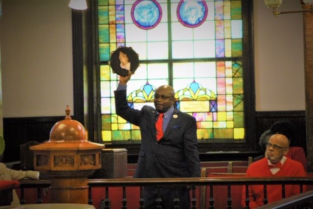 Brother Clifford Jones in suit raises tambourine inside church, stained glass in background.