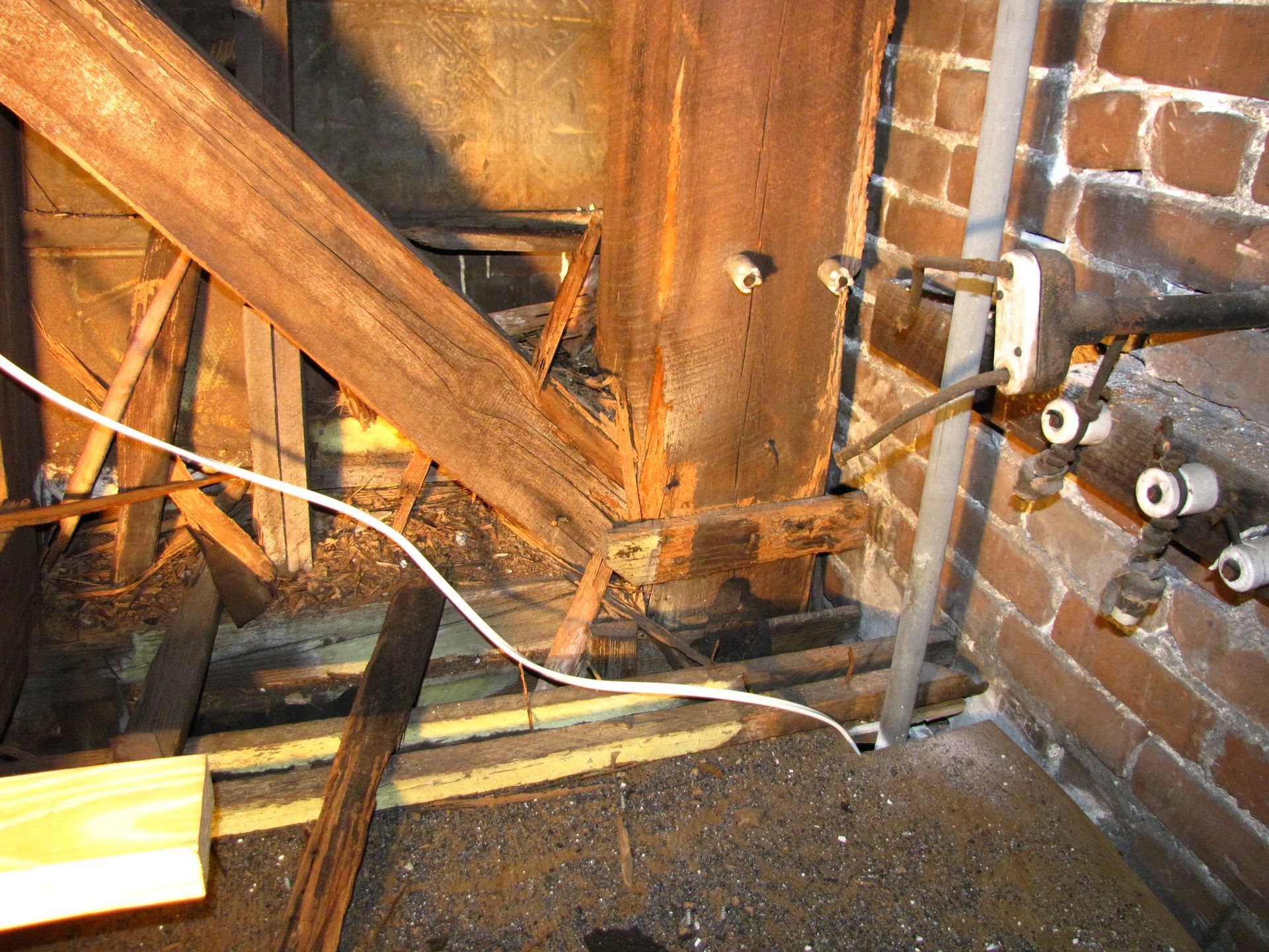 Exposed attic corner with wooden beams, brick wall, electrical wires, and a small light fixture.