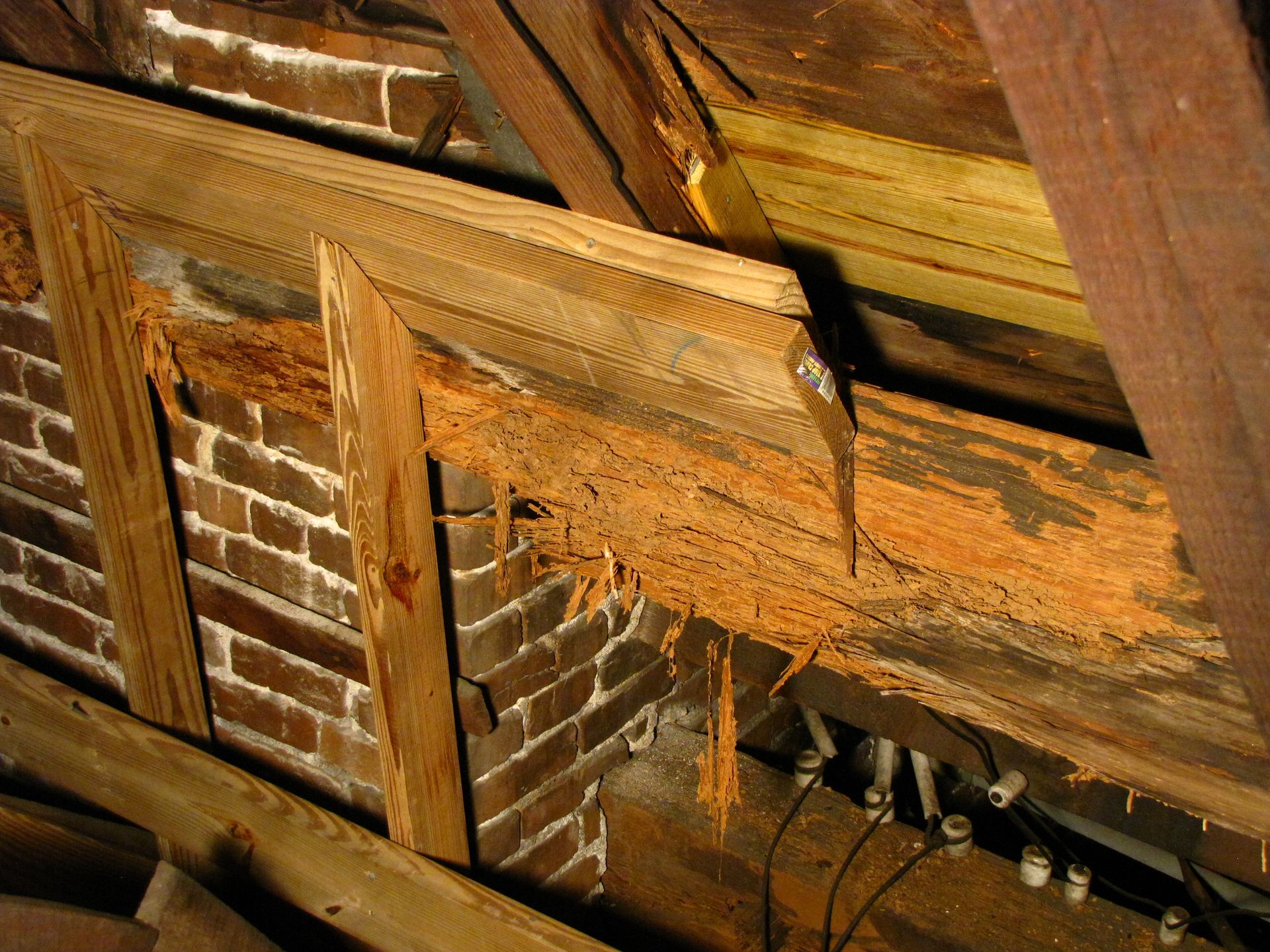 Damaged wooden roof beams and brick wall with exposed wiring inside an attic.