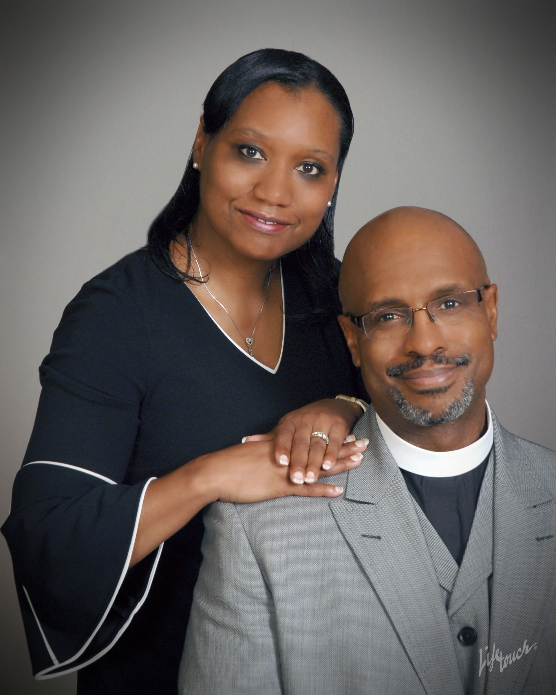 Mrs. Manning with hand on Rev. Manning's shoulder. Both smiling, in formal attire. Gray backdrop.