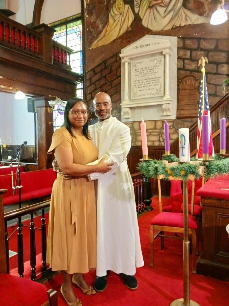 Team Manning in a church embrace near candles. Brown dress, white robe, red carpet, stained glass window.