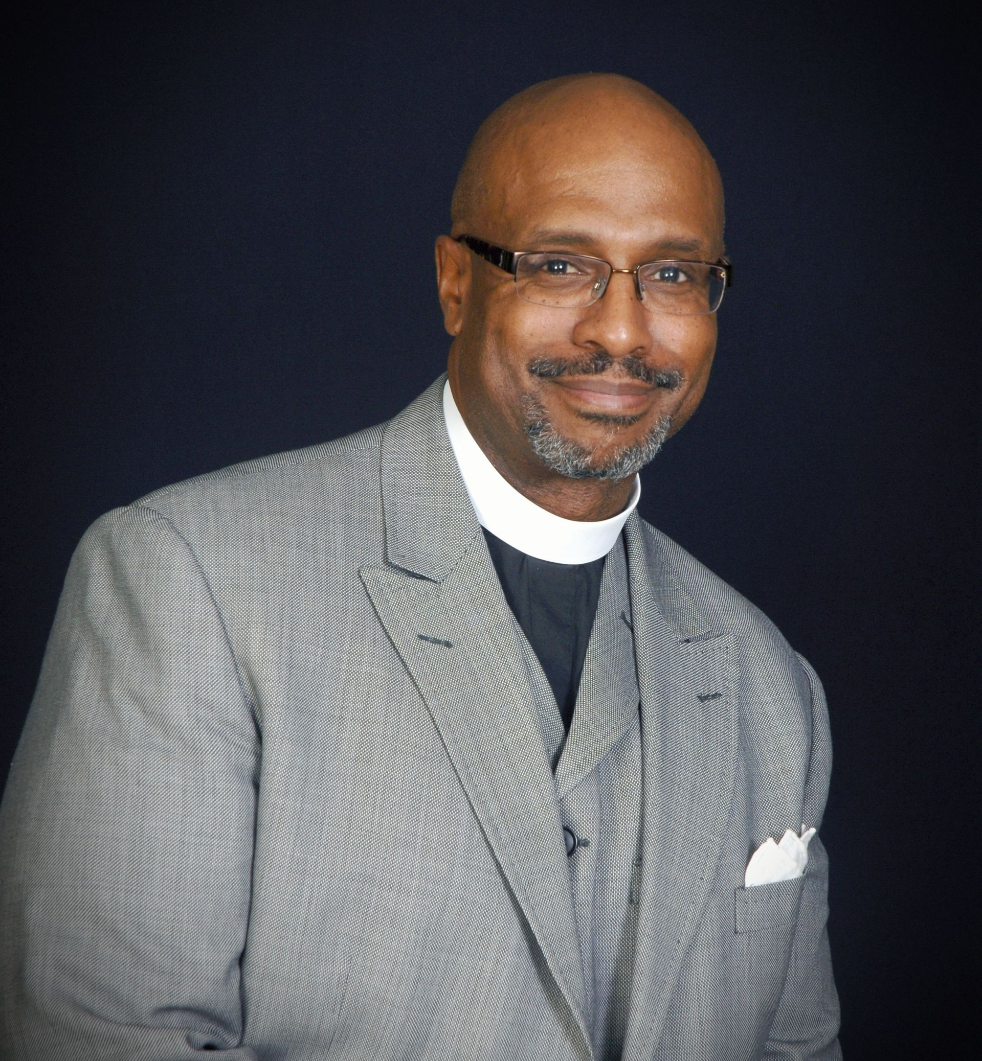 Rev. Manning in a gray suit and clerical collar, smiling, wearing glasses. Set against a dark background.