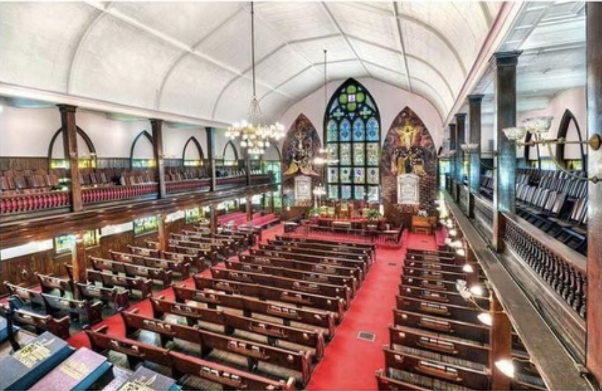 Interior of a church with wooden pews, balcony, and stained-glass windows. Red carpet and high arched ceiling.