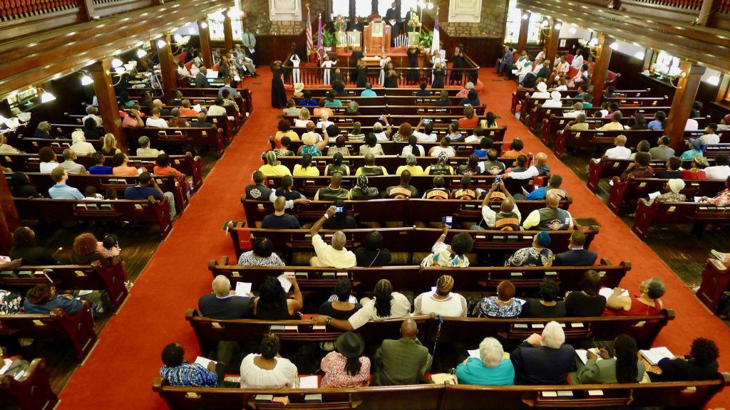 A large crowd sits in a church, facing a podium. Red carpet and dark wooden pews fill the space.
