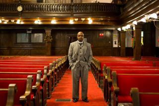 Rev. Manning in a gray suit stands in an empty church with red pews.