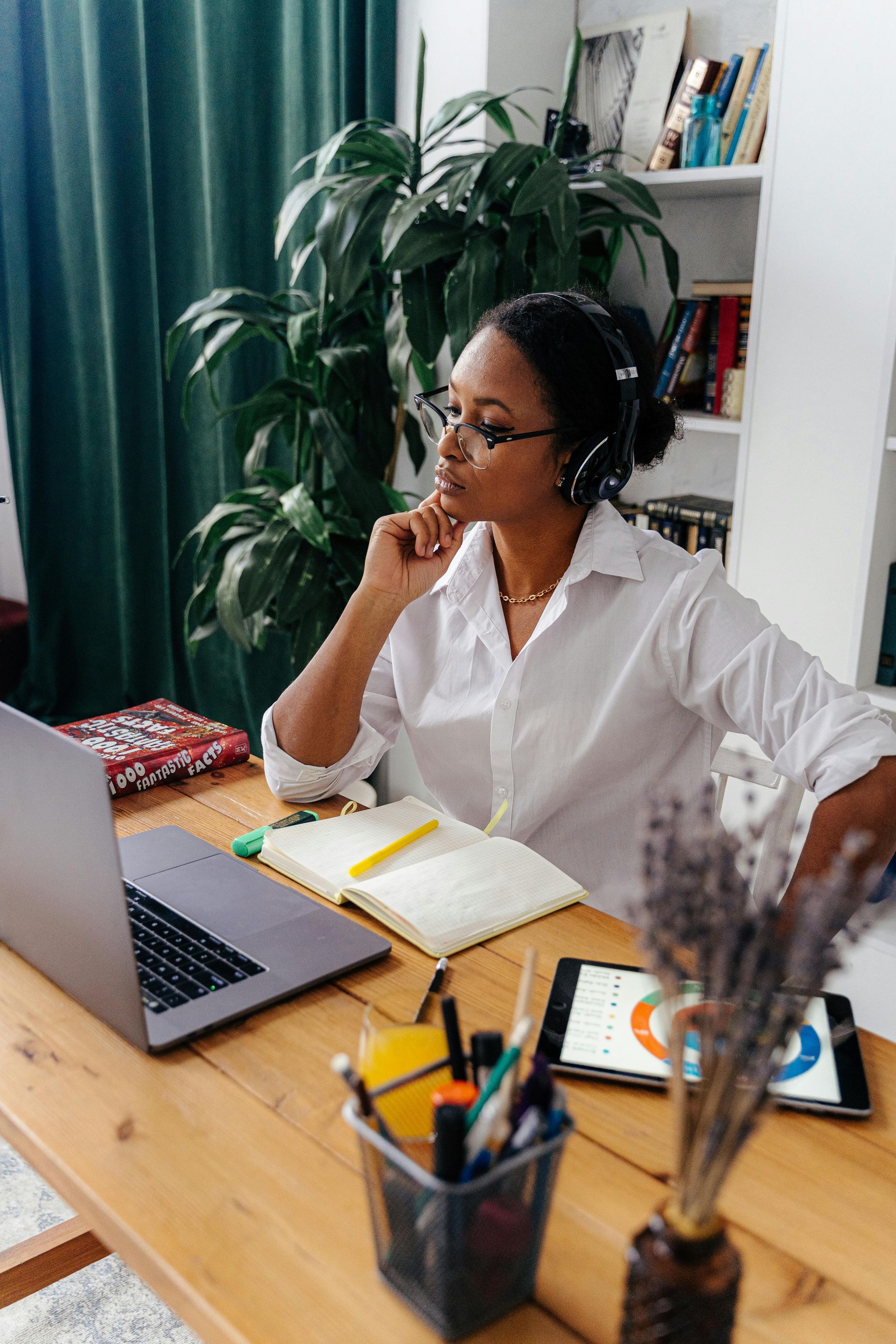 Woman at desk with laptop, tablet, and notepad, wearing headphones and glasses, looking thoughtful.