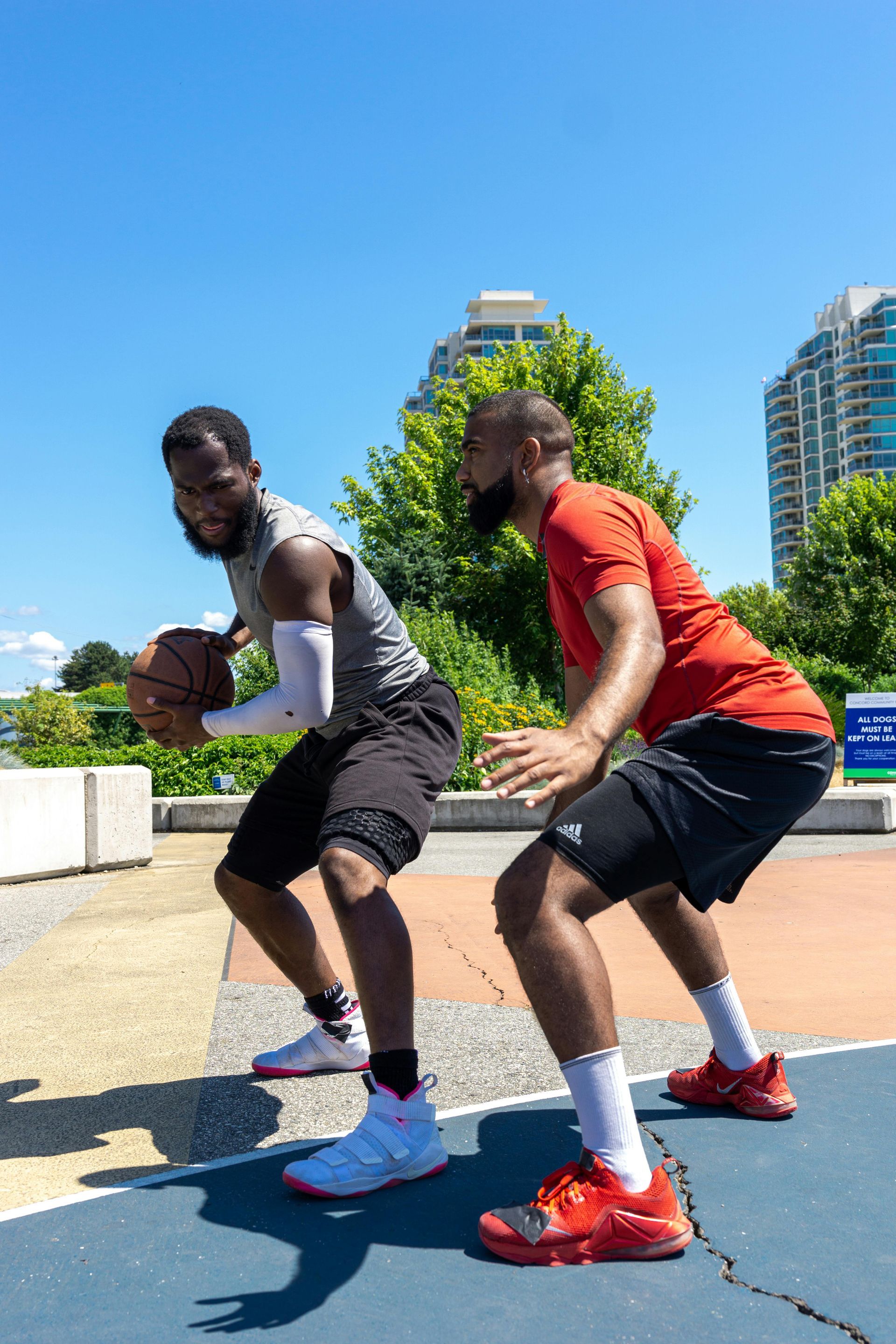 Two men playing basketball outdoors on a blue court. One dribbles, the other guards. Bright sunny day.