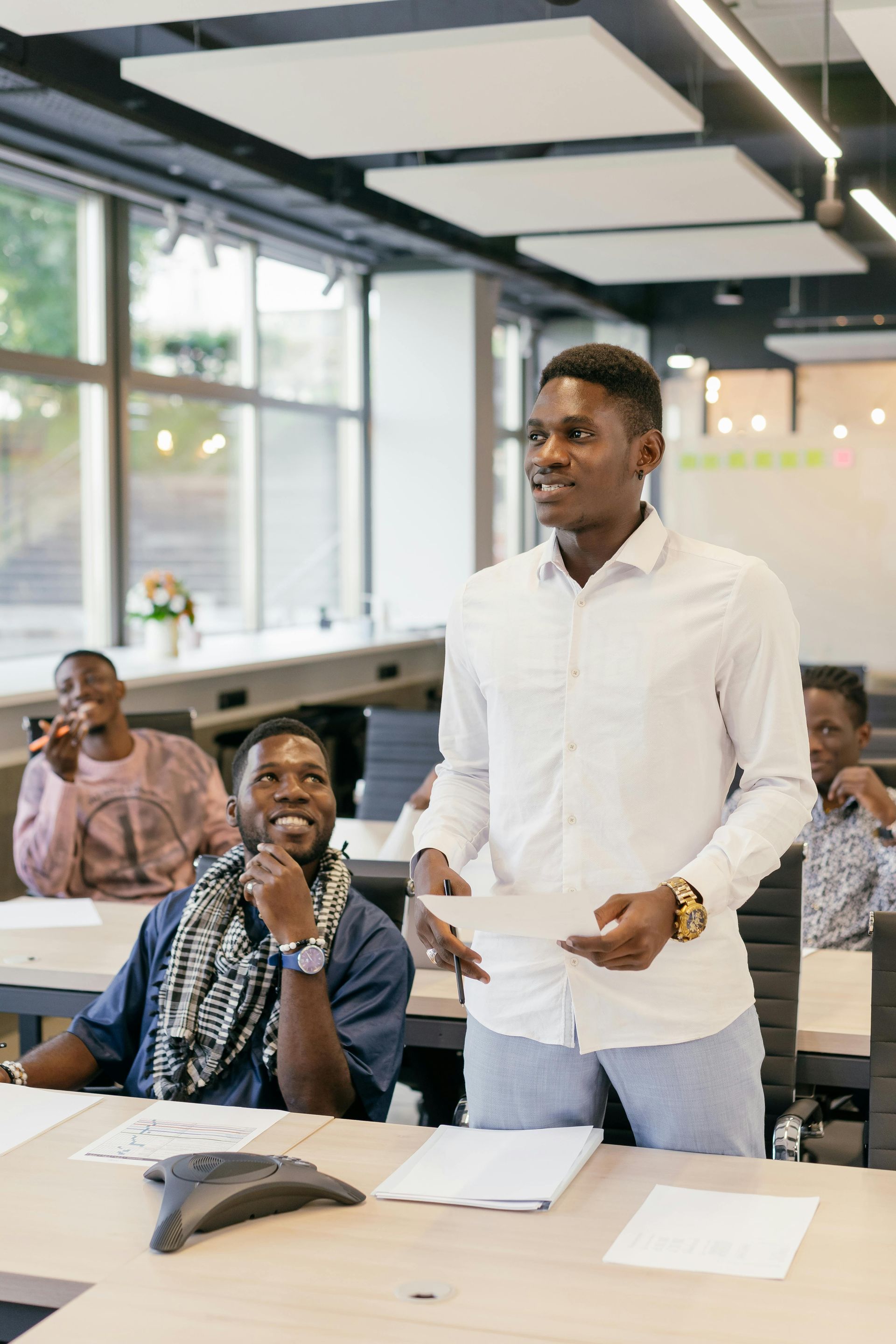 Man in white shirt presents papers in a classroom; students listen at desks.
