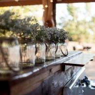 Glass jars of white flowers lined along a wooden table in a sunlit outdoor setting