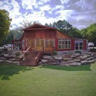 Rustic red cabin with stone landscaping and a front porch in a green yard under cloudy sky