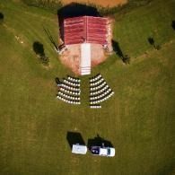 Aerial view of an outdoor ceremony setup with rows of white chairs, a red-roofed building, and a parked vehicle