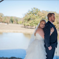 Bride in white dress and groom in navy suit smiling by a lakeside outdoors