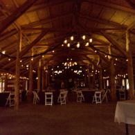 Rustic barn interior with hanging lights, wooden beams, and chairs set up for an event.