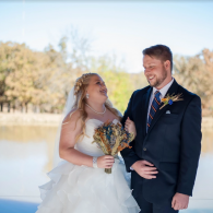 Bride and groom smiling outdoors by the water, holding a bouquet in a sunlit wedding scene