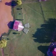 Outdoor wedding setup with chairs and an arch by a pond, and a grassy field, viewed from above