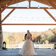 Bride and groom dancing under a wooden pavilion with string lights, overlooking a scenic autumn landscape