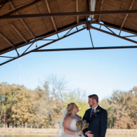 Bride and groom standing together under a wooden pavilion outdoors on a sunny day