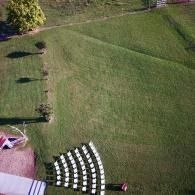 Aerial view of a large green lawn with scattered shadows and a curved row of white chairs near a patio.