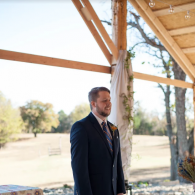 Man in a dark suit standing under a wooden pavilion outdoors, looking to the side.