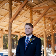 Man in a navy suit standing in a wooden pavilion with string lights and a blurred outdoor background