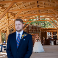 Blurred man in navy suit in foreground, bride in white gown under wooden pavilion behind him.