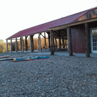 Rustic building with red metal roof, wooden porch, and gravel foreground at sunset