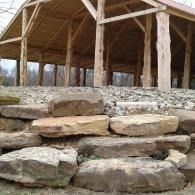 Stone retaining wall in front of a wooden pavilion with a peaked roof.