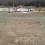 Rural yard with a small white shed and red-roofed carport near a pond, under overcast skies.