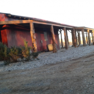 A weathered red building with a row of open supports and gravel ground at dusk