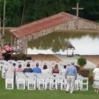 Outdoor pavilion with white chairs facing a landscaped area and cross, surrounded by trees.