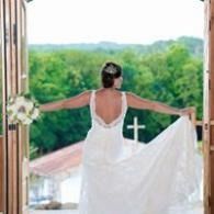 Woman in a white dress standing in a doorway with arms outstretched, overlooking green hills and a cross.